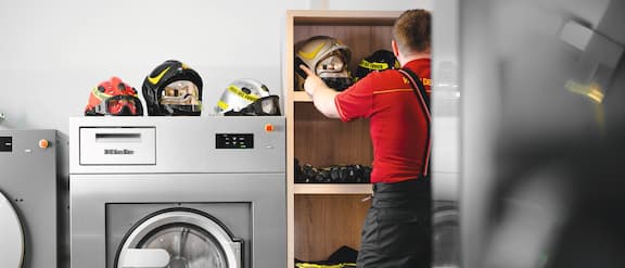 A fireman stores various helmets in a cupboard. Some helmets are also resting on top of the Miele Benchmark Performance Plus washing machine next to him.