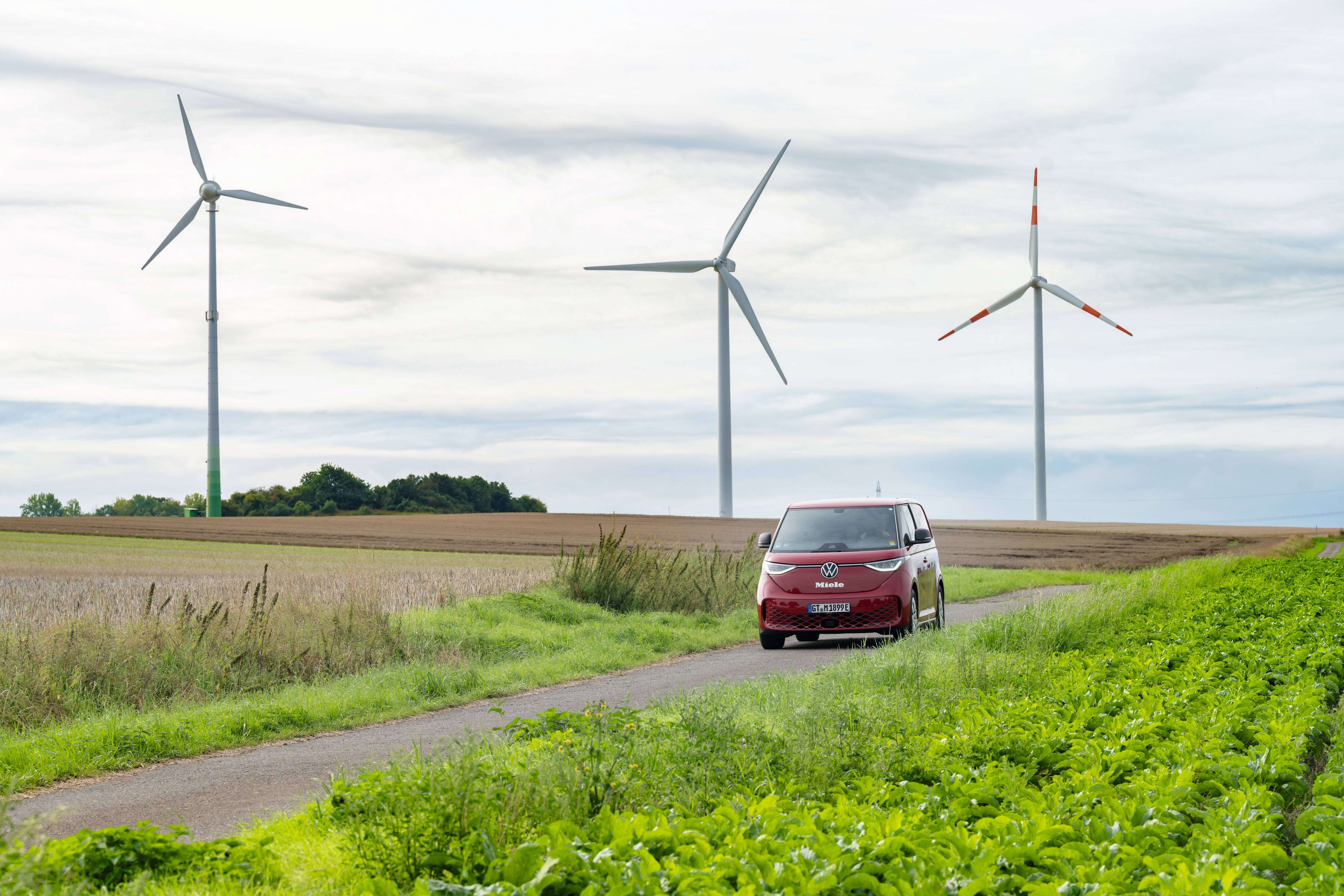 Czerwona furgonetka Volkswagena na wiejskiej drodze z turbinami wiatrowymi w tle, symbolizująca ekologiczny transport i energię odnawialną