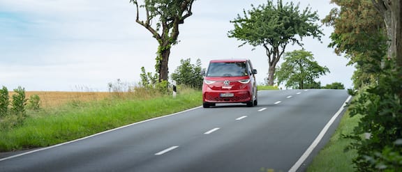 The red Miele Service VW ID Buzz drives along a country road. There are a few trees along the side of the road.
