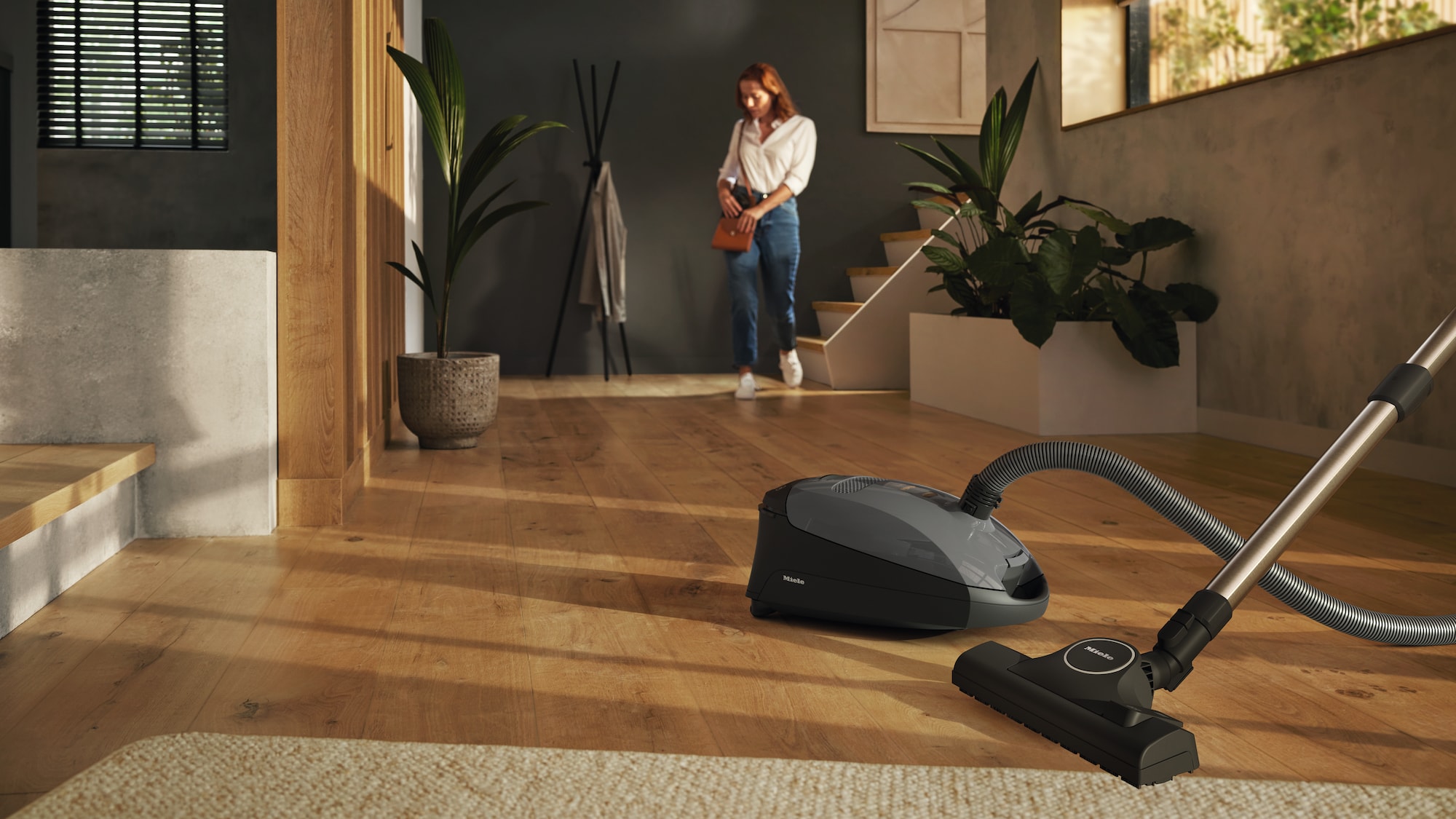 Grey canister vacuum sitting on a wooden floor of a house with a woman walking down the stairs in the background