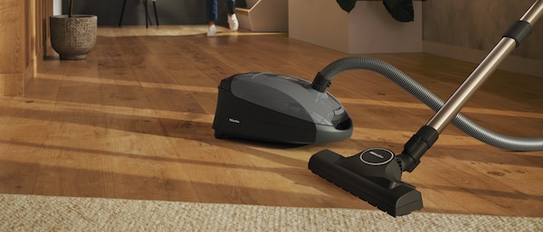 Grey bagged canister vacuum on wood floor of house with woman walking in the background towards the vacuum