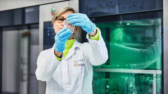 Eine Labormitarbeiterin steht vor Laborspülern, die grün leuchten und prüft die Sauberkeit eines Laborglases.