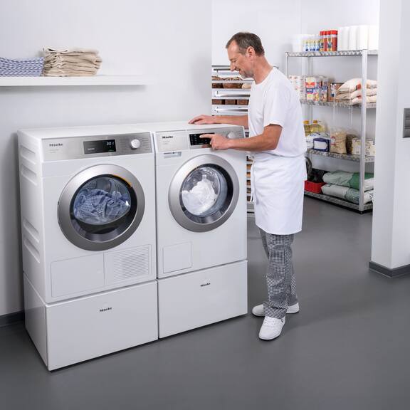 En frisør står foran Mieles SmartBiz PWM 1108-vaskemaskinen og indstiller et vaskeprogram. Ved siden af PWM 1108 står Mieles SmartBiz PDR 1108 HP-varmepumpetørretumbler