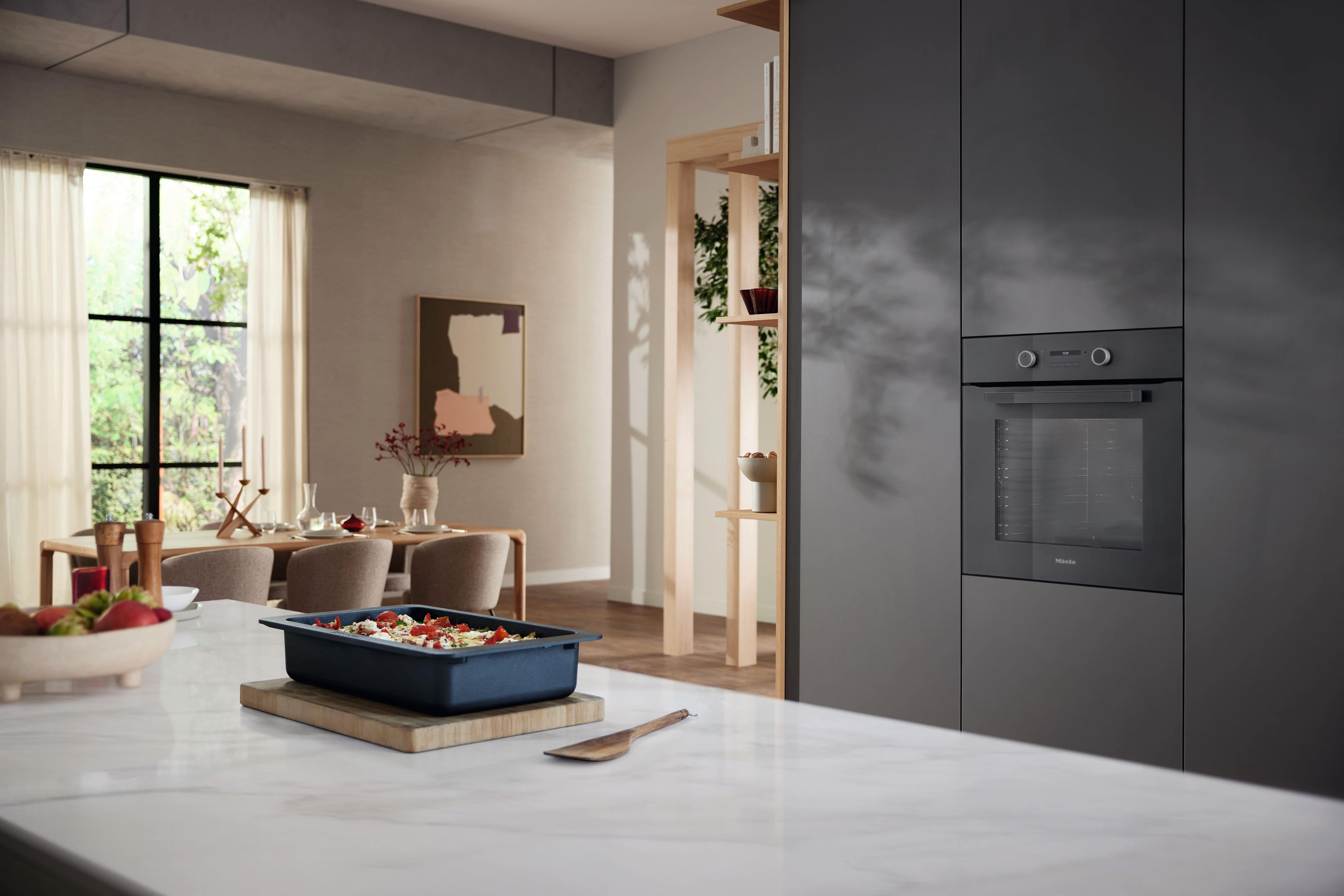 Image of kitchen with food in a tray on top of a wooden board with a wooden spoon next to it, there is a kitchen table and chairs and a black Miele built in oven in the background