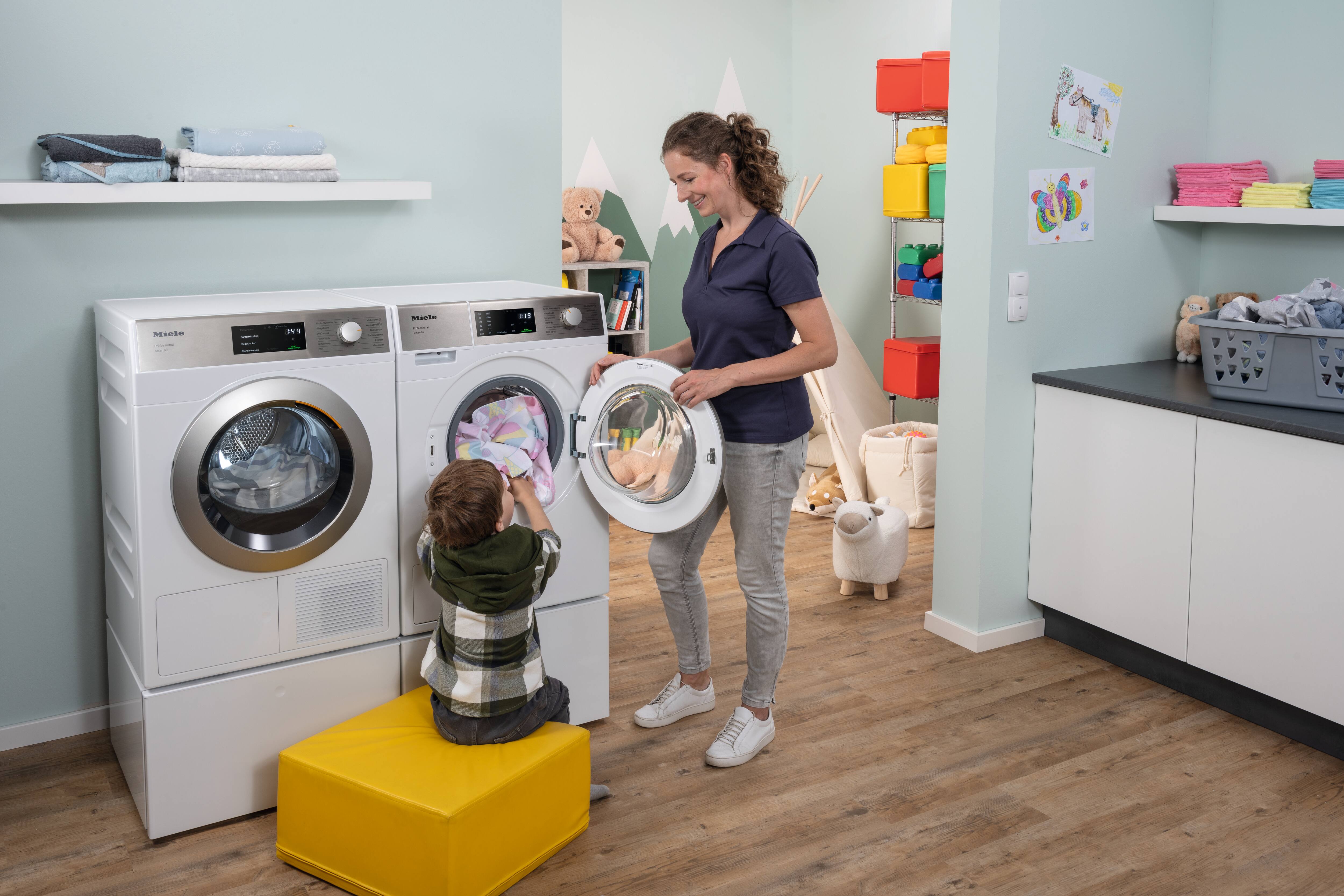 En pædagog bærer en grå vasketøjskurv fyldt med farverige hagesmække og smiler til en dreng, der sidder i et legetelt. Mieles SmartBiz PWM 1108-vaskemaskine og PDR 1108 HP-varmepumpetørretumbler står til venstre for pædagogen.