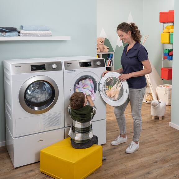 En pædagog bærer en grå vasketøjskurv fyldt med farverige hagesmække og smiler til en dreng, der sidder i et legetelt. Mieles SmartBiz PWM 1108-vaskemaskine og PDR 1108 HP-varmepumpetørretumbler står til venstre for pædagogen.