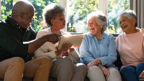 Drei glückliche verschiedene ältere Frau und ein männlicher Freund sitzen auf Sofa und verwenden Tablet