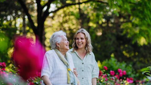 Eine ältere Dame mit einem Rollator geht mit einer jungen Dame im Außenbereich eines Pflegeheims spazieren