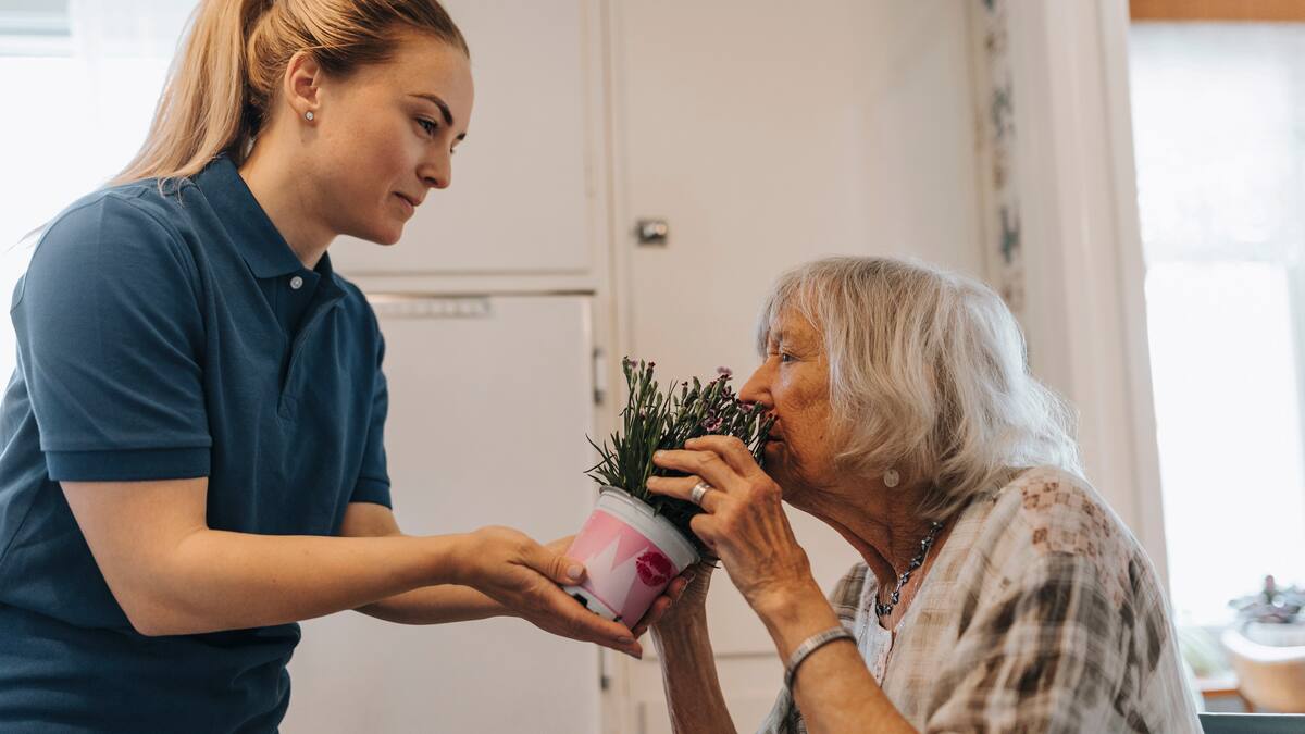 Une dame âgée sent une plante en pot tenue par une soignante
