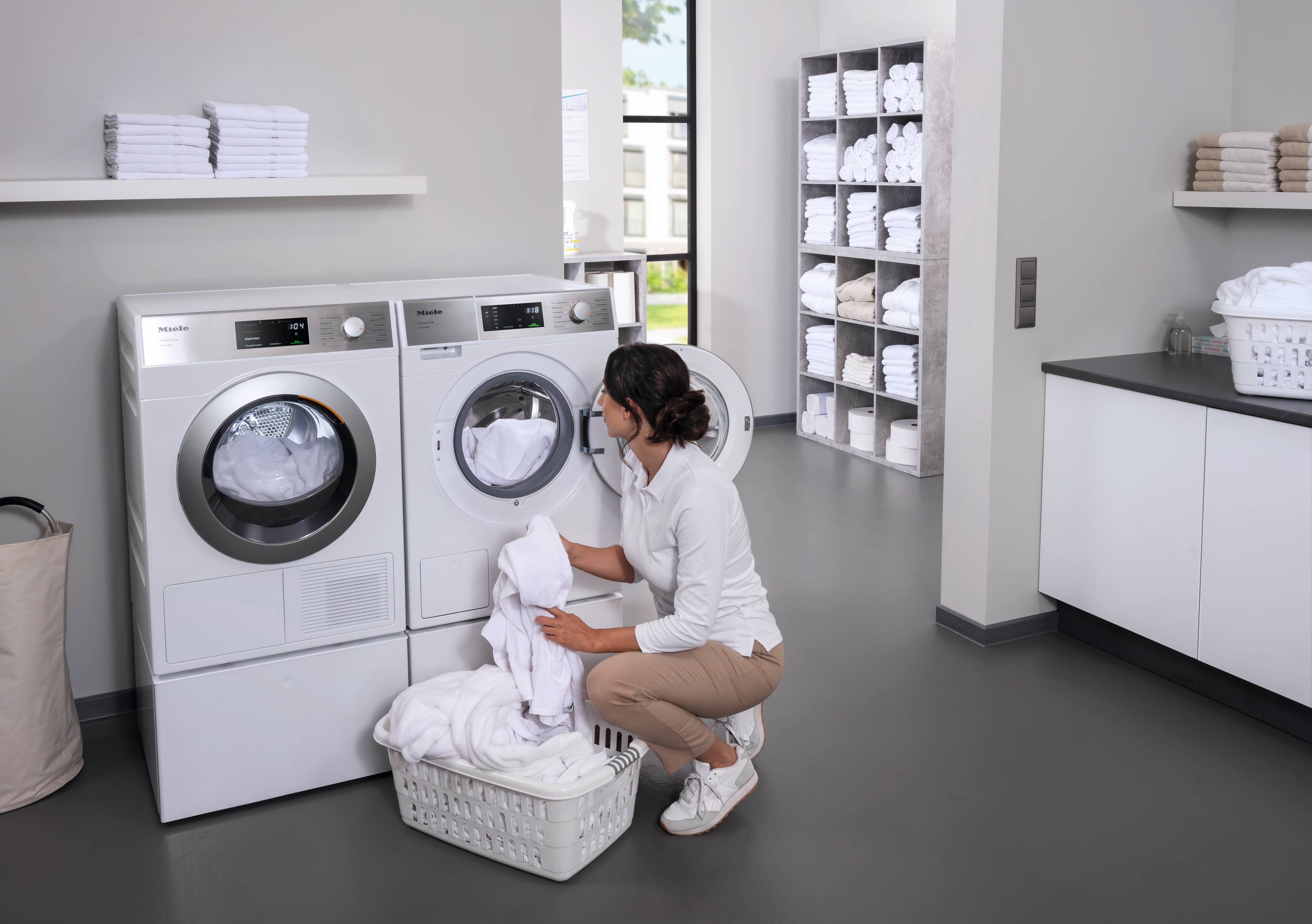 En kosmetolog fylder en Miele SmartBiz PWM 1108-vaskemaskine med hvide badekåber. Ved siden af vaskemaskinen står Mieles SmartBiz PDR 1108 HP-varmepumpetørretumbler.