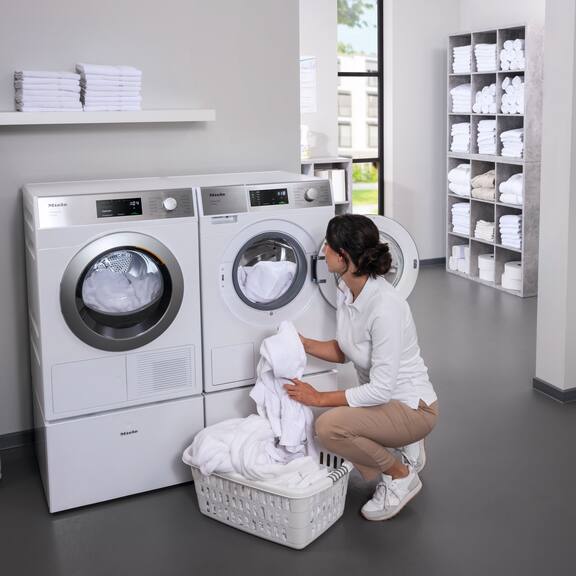 En kosmetolog fylder en Miele SmartBiz PWM 1108-vaskemaskine med hvide badekåber. Ved siden af vaskemaskinen står Mieles SmartBiz PDR 1108 HP-varmepumpetørretumbler.