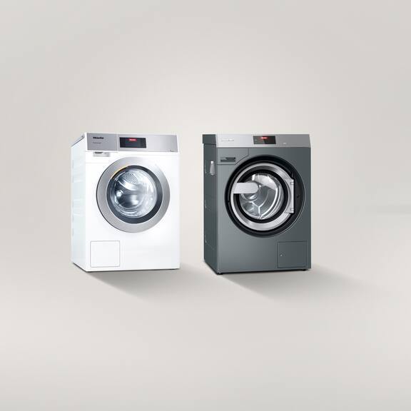 Een Kleine Reuzen Performance Plus wasmachine staat links naast een Benchmark Performance wasmachine tegen een grijze achtergrond