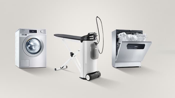 Von links nach rechts stehen eine Kleine Riesen Performance Waschmaschine, ein PIB100 Dampfbügelsystem sowie eine halb geöffnete MasterLine Frischwasser-Spülmaschine nebeneiner vor einem grauen Hintergrund