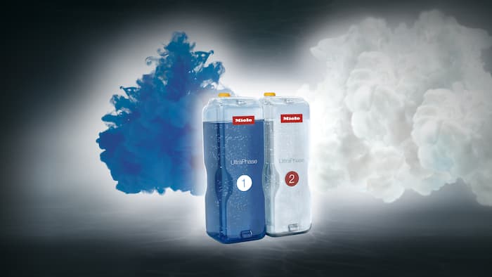 Kartuschen der Miele TwinDosWaschmittel UltraPhase 1 und UltaPhase 2 stehen vor einem grauen Hintergrund. Neben den Kartuschen entweichen eine blaue und eine weiße Farbwolke.