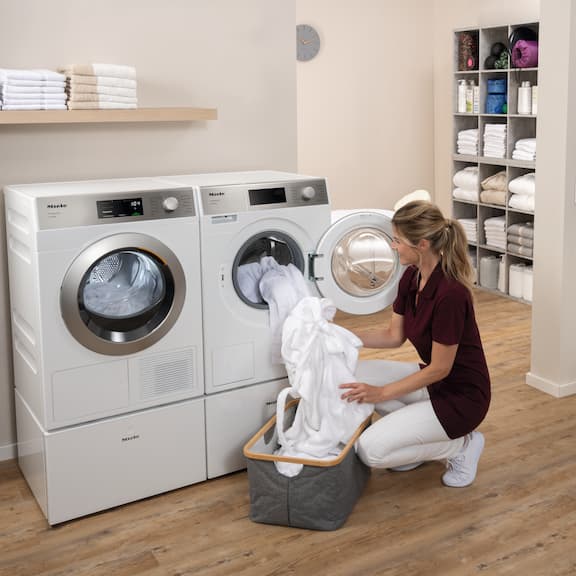 En kosmetolog fyller en Miele SmartBiz-tvättmaskin PWM 1108 med vita badrockar. Bredvid tvättmaskinen står Mieles SmartBiz-värmepumpstumlare PDR 1108 HP.