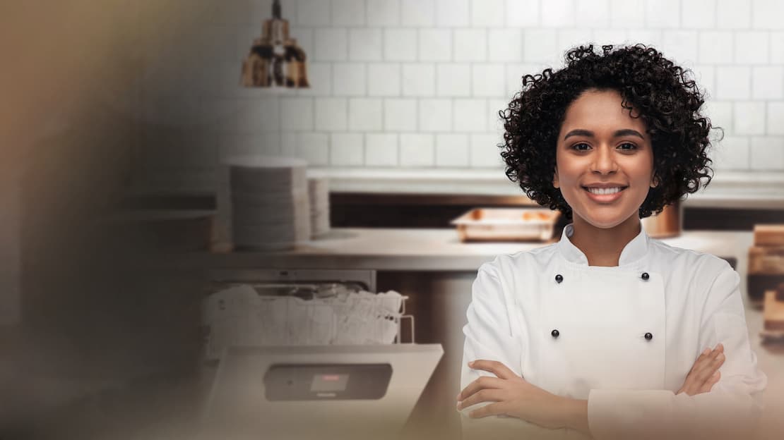 A female chef is standing smiling in front of the commercial Miele MasterLine dishwasher in a restaurant kitchen.