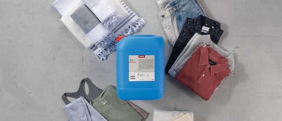 A media canister containing ProCare Tex detergent is placed on a grey floor. Various items of clothing are artistically draped in a circle around the canister.