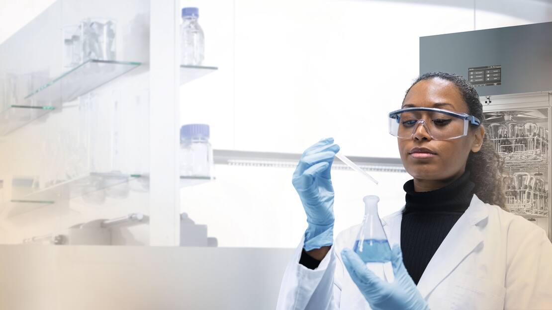 Eine Labormitarbeiterin hält eine Pipette und ein Laborglas und steht vor einem Laborspüler in einer weißen Laborumgebung.