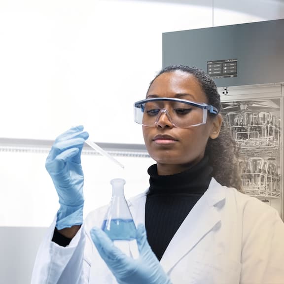 A lab technician holding a pipette and an item of laboratory glassware, and standing in front of a laboratory washer in a white laboratory environment.