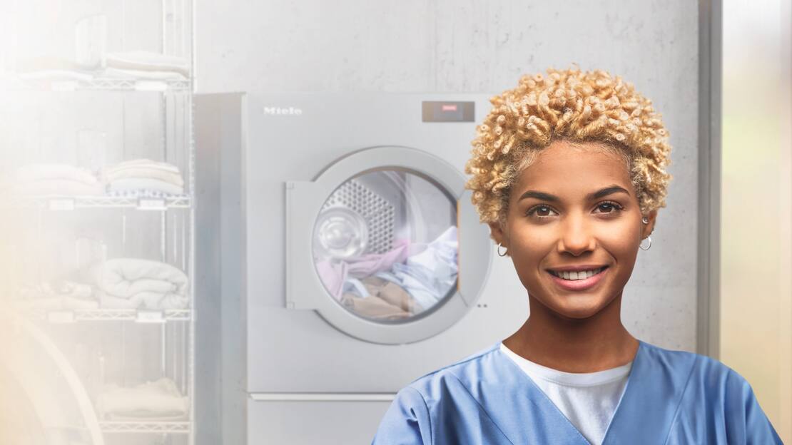A care worker is standing smiling in front of a Miele Benchmark Performance Plus dryer and a shelf stocked with laundry.
