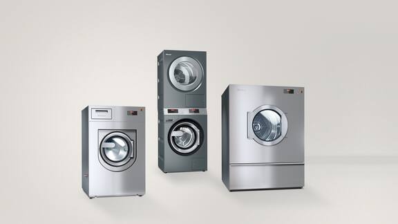 Von links nach rechts stehen eine Kleine Riesen Performance Waschmaschine, ein PIB100 Dampfbügelsystem sowie eine halb geöffnete MasterLine Frischwasser-Spülmaschine nebeneiner vor einem grauen Hintergrund