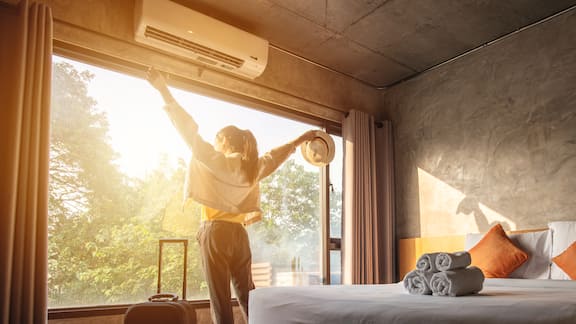 A young woman stands at the window of her hotel room with her suitcase and looks out. She raises her arms in the air and holds a hat in her hand. The sun is shining through the window.
