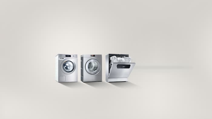Fra venstre mod højre står en kompakt Performance-vaskemaskine, et PIB100 dampstrygesystem samt en halvåben MasterLine friskvandsopvaskemaskine ved siden a hinanden foran en grå baggrund