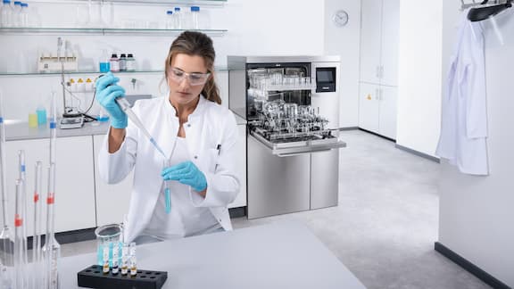 Technik laboratoryjny przeprowadza analizę, napełniając probówkę cieczą za pomocą pipety. W tle znajduje się otwarta myjnia laboratoryjna PLW 8636.