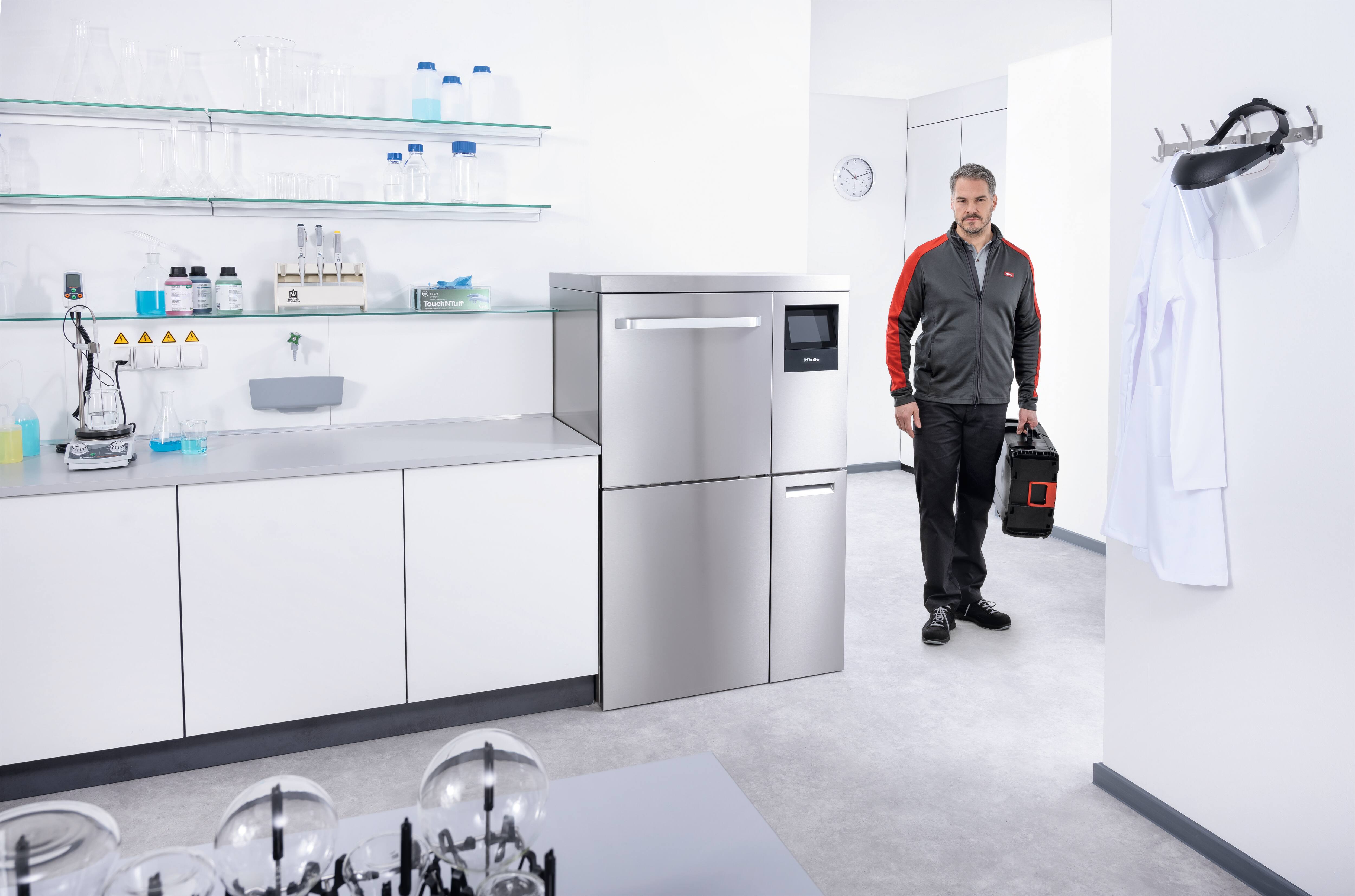 Ein Miele Servicetechniker geht in ein Labor mit einem Werkzeugkoffer in der Hand. Neben ihm steht ein Laborspüler PLW 8636.