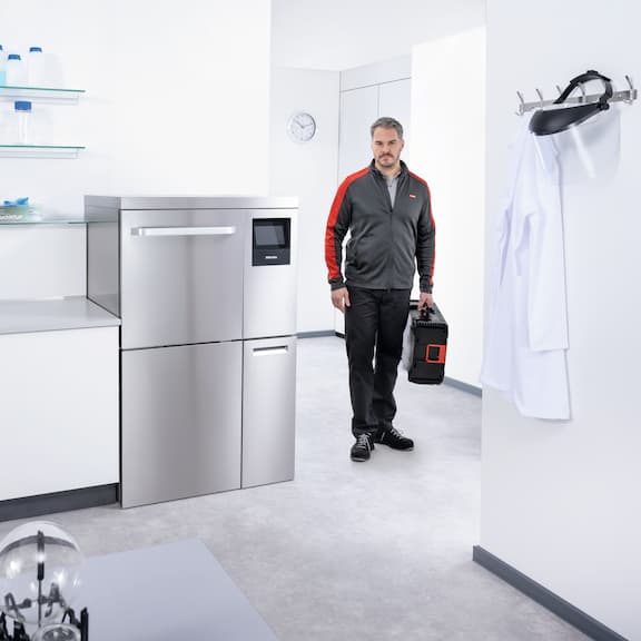 Serwisant Miele wchodzi do laboratorium ze skrzynką narzędziową. Obok niego znajduje się myjnia laboratoryjna PLW 8636.