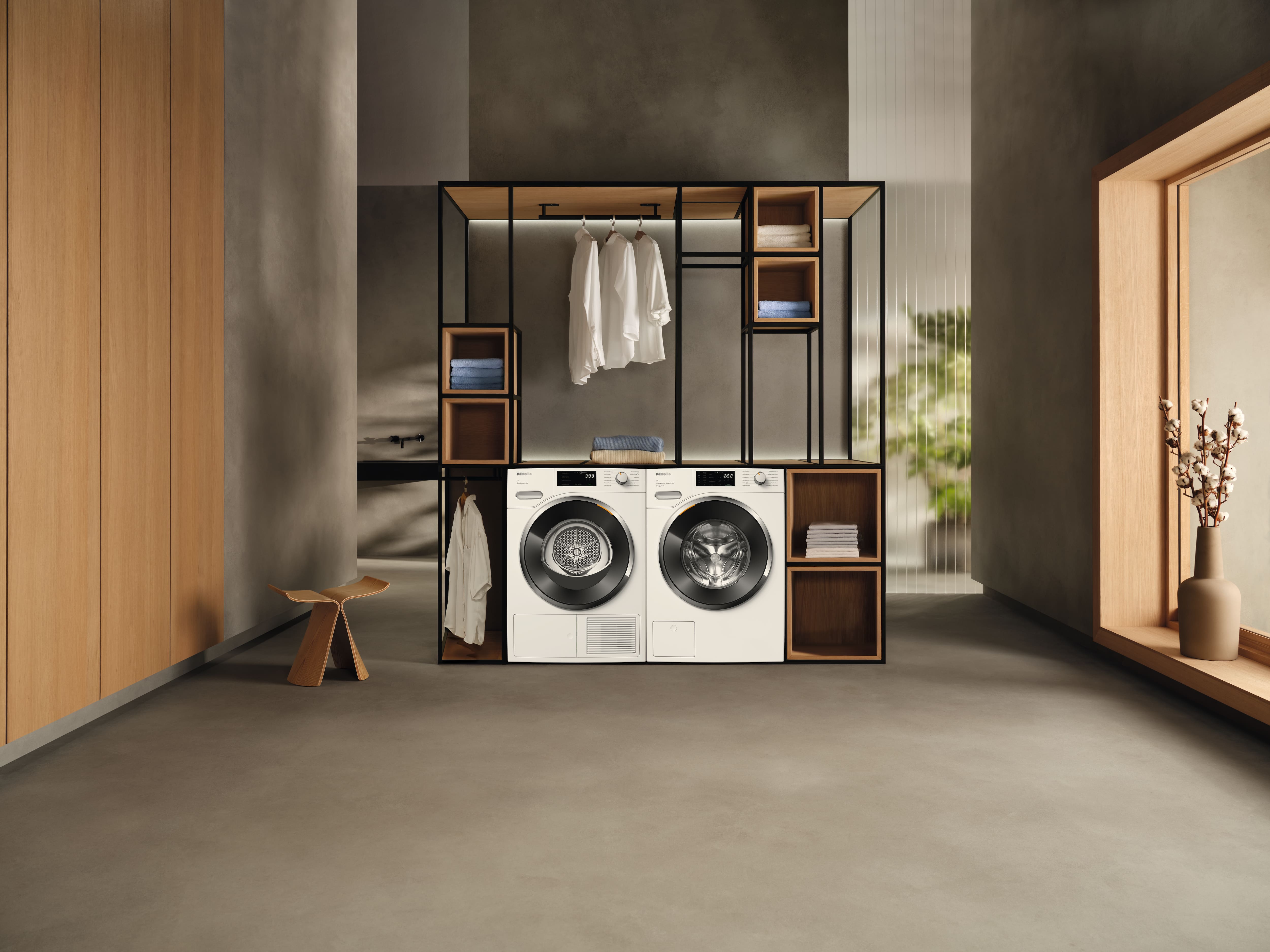 A minimalist laundry setup with a washer and dryer beneath an open black-framed shelving unit holding folded clothes, hanging garments, and storage cubbies, set in a modern room with neutral tones and large windows. A minimalist laundry setup with a washer and dryer beneath an open black-framed shelving unit holding folded clothes, hanging garments, and storage cubbies, set in a modern room with neutral tones and large windows.
