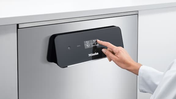 En finger trykker på et sort touchdisplay med ikoner, som er en del af en laboratorieopvaskemaskine i rustfrit stål.