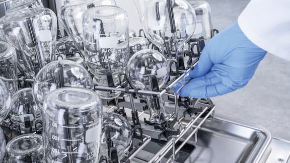 En hånd skubber et EasyLoad-modul fyldt med laboratorieglas ned i en underkurv. Laboratorieglassene er i forskellige størrelser og er en blanding af rundkolber, flasker og erlenmeyerkolber.