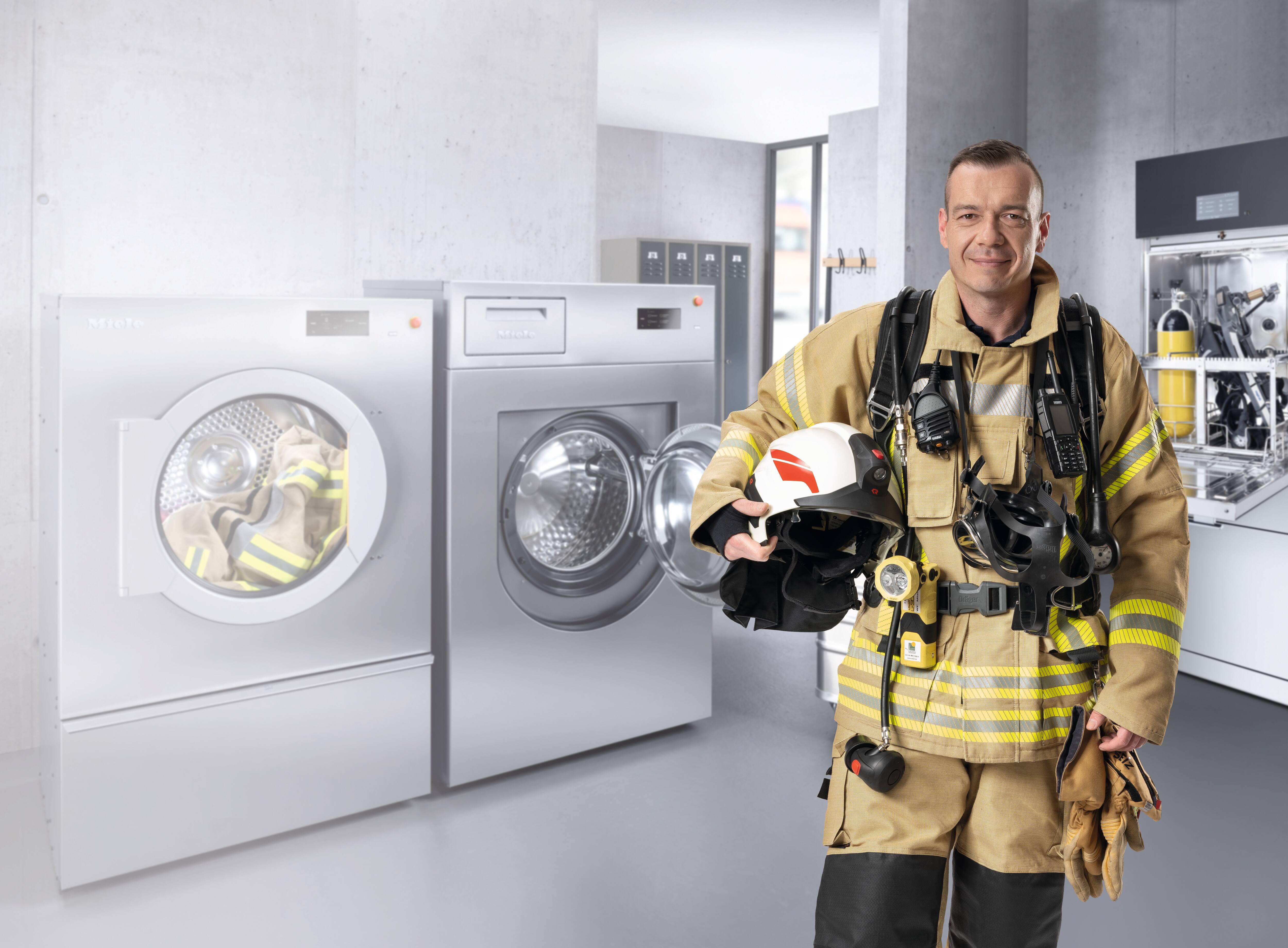Ein Feuerwehrmann in vollstäniger PSA guckt lächelnd in die Kamera. Im Hintergrund stehen die Miele Benchmark Performance Plus Waschmaschine und Trockner, sowie die PLW 7111 Safety.