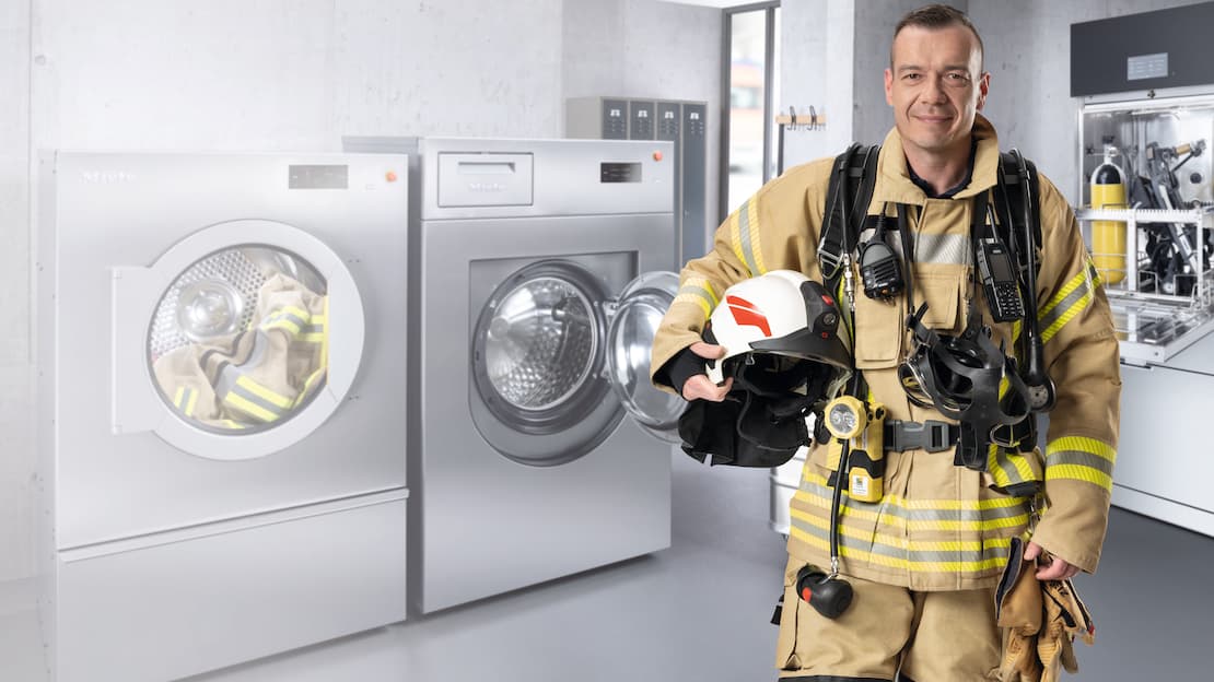 A firefighter in full personal protective equipment looks into the camera with a smile. A Miele Benchmark Performance Plus washing machine and dryer, and the PLW 7111 Safety, can be seen in the background.