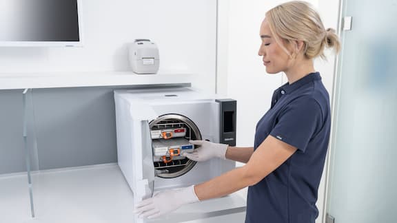 Une assistante dentaire dans la salle de préparation ouvre un stérilisateur Cube Miele avec des conteneurs de stérilisation EasyStore.