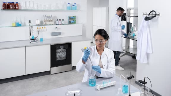 En laborant sidder ved et bord og udfører en laboratorieanalyse. En anden laboratorietekniker doserer væske i baggrunden, og en ExpertLine-laboratorieopvaskemaskine med glasdør er indbygget i laboratoriet som en underbygningsmodel.