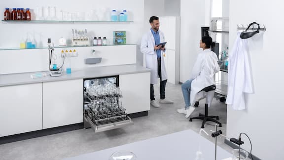 A ExpertLine laboratory washer with glass door built into a run of laboratory units. In the background, a lab technician is fetching new buffer solution from a storage cabinet.