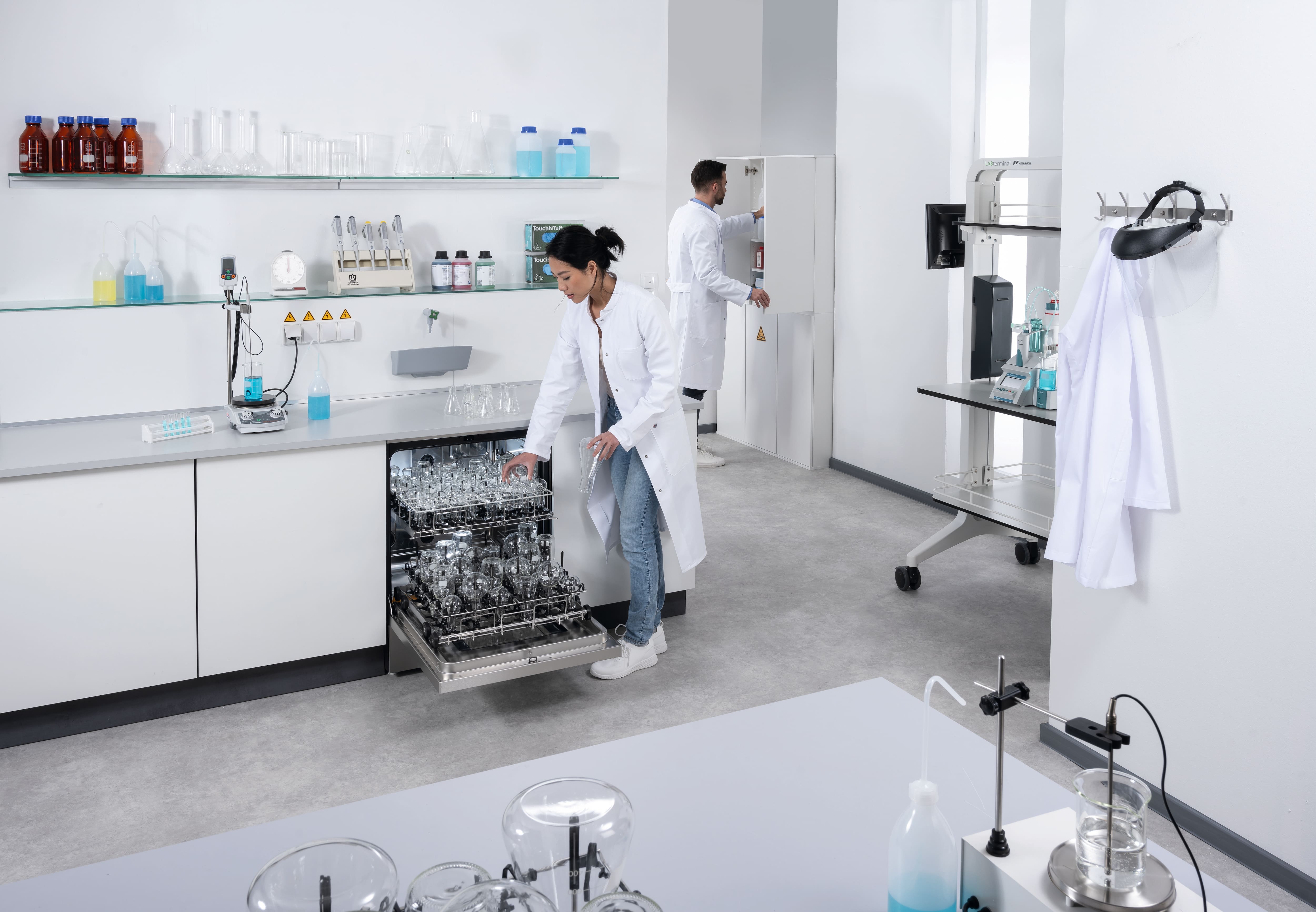 Labormitarbeiterin im Laborkittelbestückt einen geöffneten Untertischlaborspüler mit Laborglas.