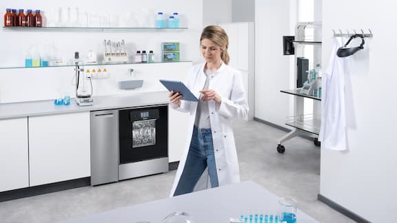 Ein Labormitarbeiter sitzt mit einem Laptop an einem Tisch vor einer Laborzeile mit einem ExpertLine Laborspüler. Auf dem Laptopscreen ist Miele MOVE Connect zu sehen mit einem Programmablauf.