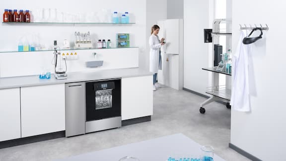 En 90 cm ExpertLine-laboratorieopvaskemaskine med glasdør indbygget i et arbejdsbord. I baggrunden henter en laboratorietekniker ny bufferopløsning fra et opbevaringsskab.