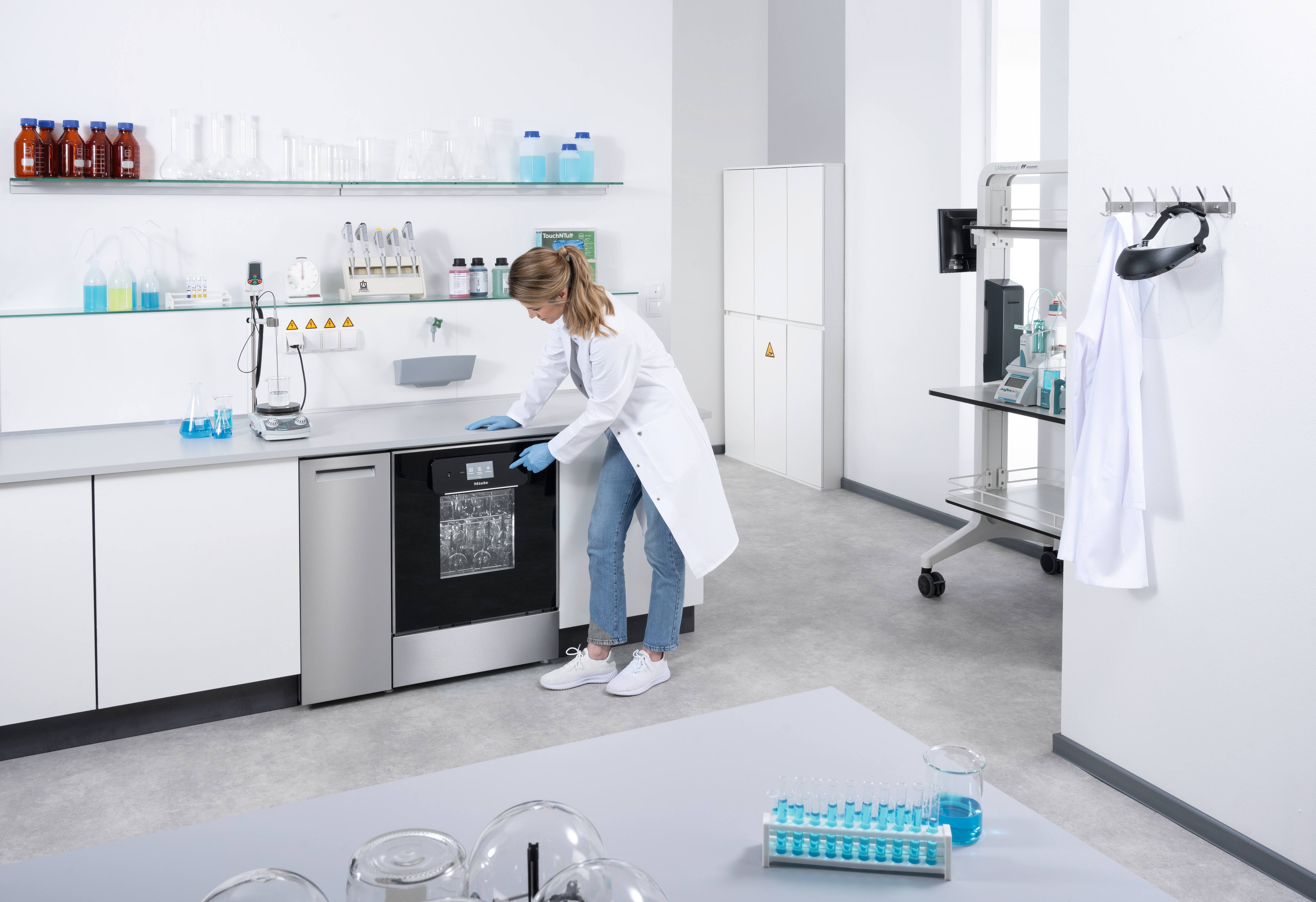 Eine Labormitarbeiterin bedient das Touch Display eines Untertisch-Laborspülers mit Glastür, um ein Programm zu starten.