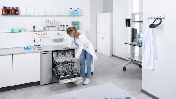 A lab technician putting instruments into lab dishwasherratory analysis. Another lab technician is dispensing liquid in the background and an ExpertLine laboratory washer with glass door is built into the laboratory as an undercounter unit.