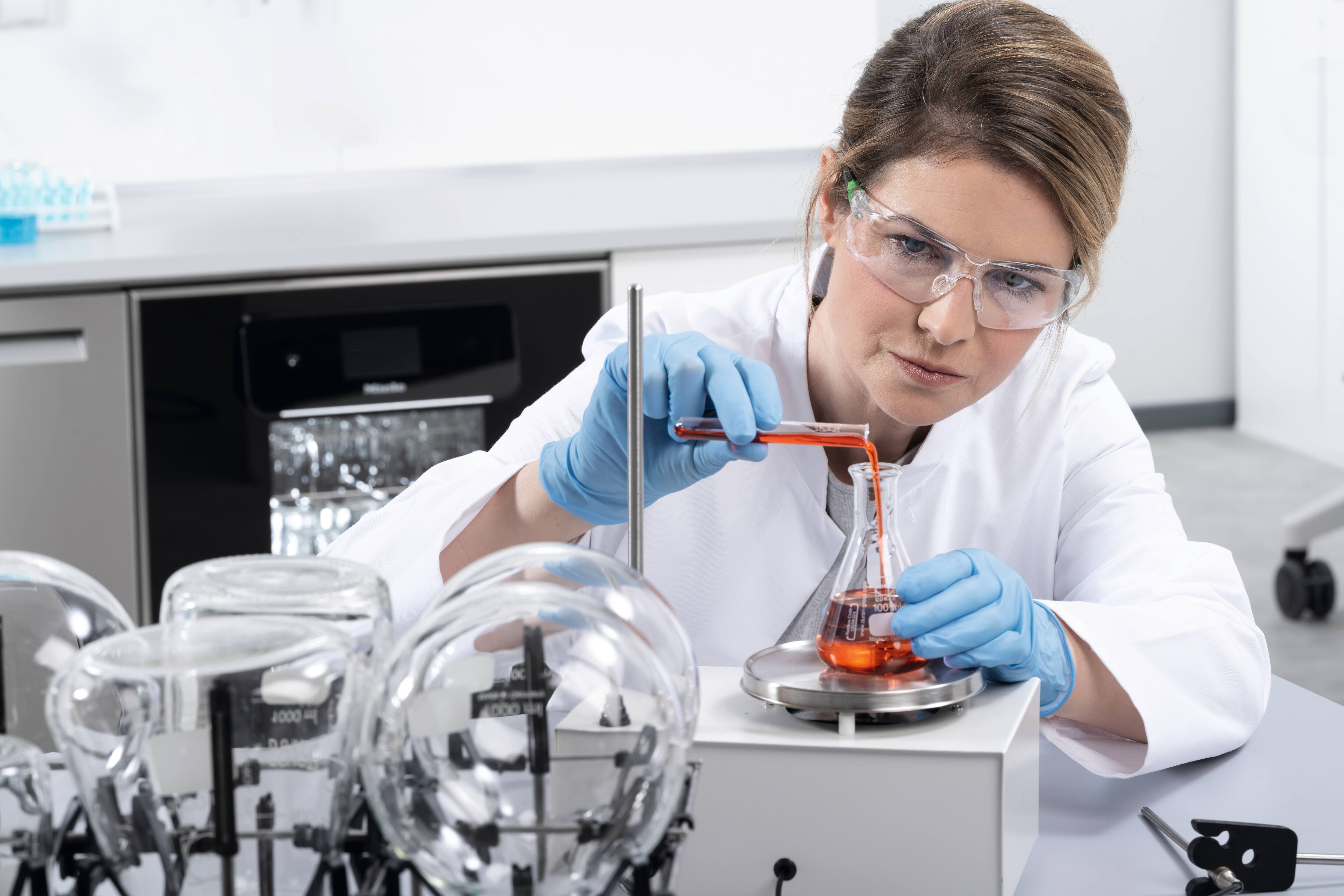 A laboratory assistant pours red liquid from a test tube into an Erlenmeyer flask.