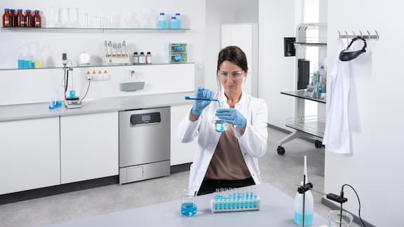 En laborant fylder væske fra et reagensglas over i en rundkolbe. I baggrunden er en ExpertLine-laboratorieopvaskemaskine i rustfrit stål indbygget i et arbejdsbord.