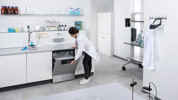Eine Labormitarbeiterin öffnet einen Laborspüler mit einwandfrei getrocknetem Laborglas.
