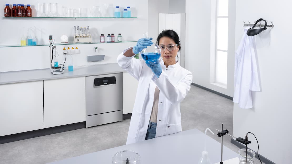 En laboratorietekniker står foran et arbejdsbord med laboratorieenheder. ExploreLine PLW 8604-laboratorieopvaskemaskine i rustfrit stål kan ses i baggrunden.