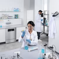 En ExploreLine-laboratorieopvaskemaskine i rustfrit stål er indbygget i et arbejdsbord. To laboratorieteknikere arbejder på forskellige laboratoriemaskiner - en kvinde i forgrunden pipetterer blå væske i reagensglas, og en mand bruger en elektrisk doseringshjælp.
