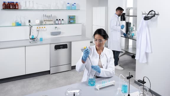 En ExploreLine-laboratorieopvaskemaskine i rustfrit stål er indbygget i et arbejdsbord. To laboratorieteknikere arbejder på forskellige laboratoriemaskiner - en kvinde i forgrunden pipetterer blå væske i reagensglas, og en mand bruger en elektrisk doseringshjælp.