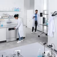 En laboratorietekniker betjener displayet på en laboratorieopvaskemaskine i rustfrit stål fra ExploreLine, mens en anden laboratorietekniker i baggrunden indtaster data på en skærm.